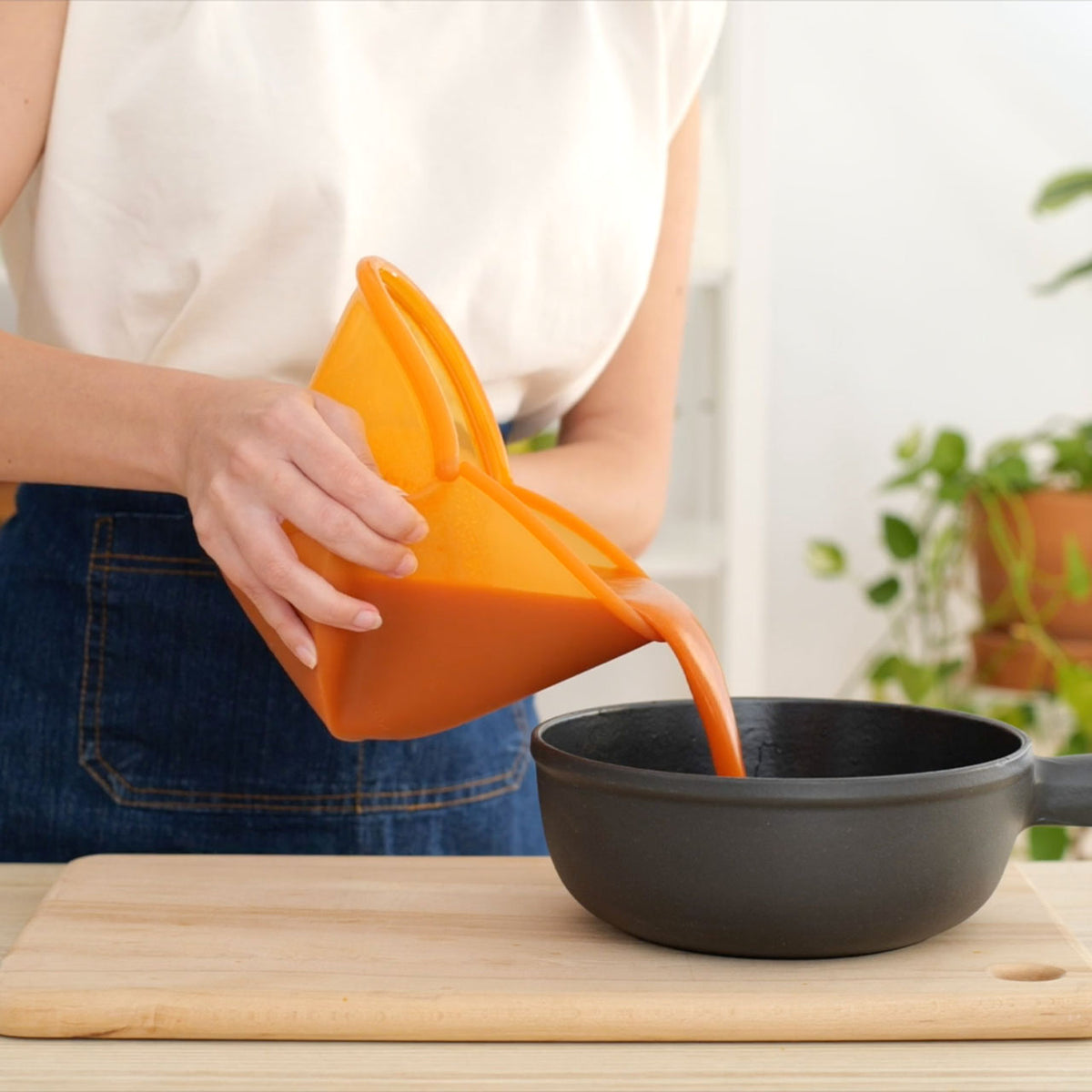 Person gießt orange Flüssigkeit aus einem Food Huggers Silikon-Frischhaltebeutel in eine schwarze Pfanne