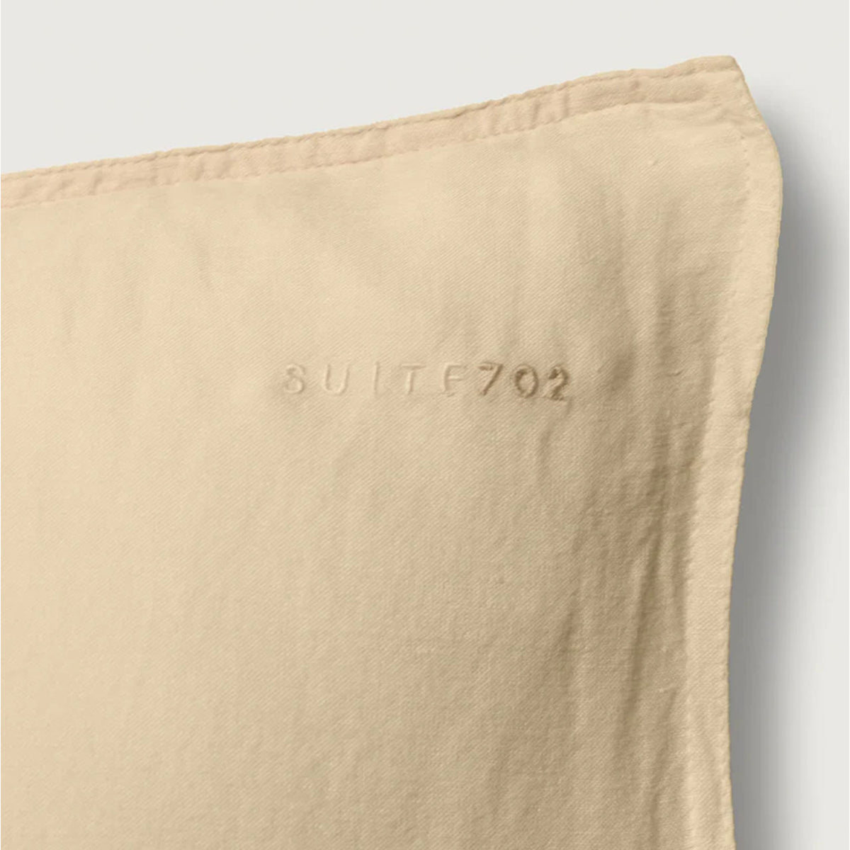 Nahaufnahme der Ecke des Suite702 Leinen-Mix Kissenbezugs in Creamy Yellow mit eingeprägtem „Suite702“-Schriftzug