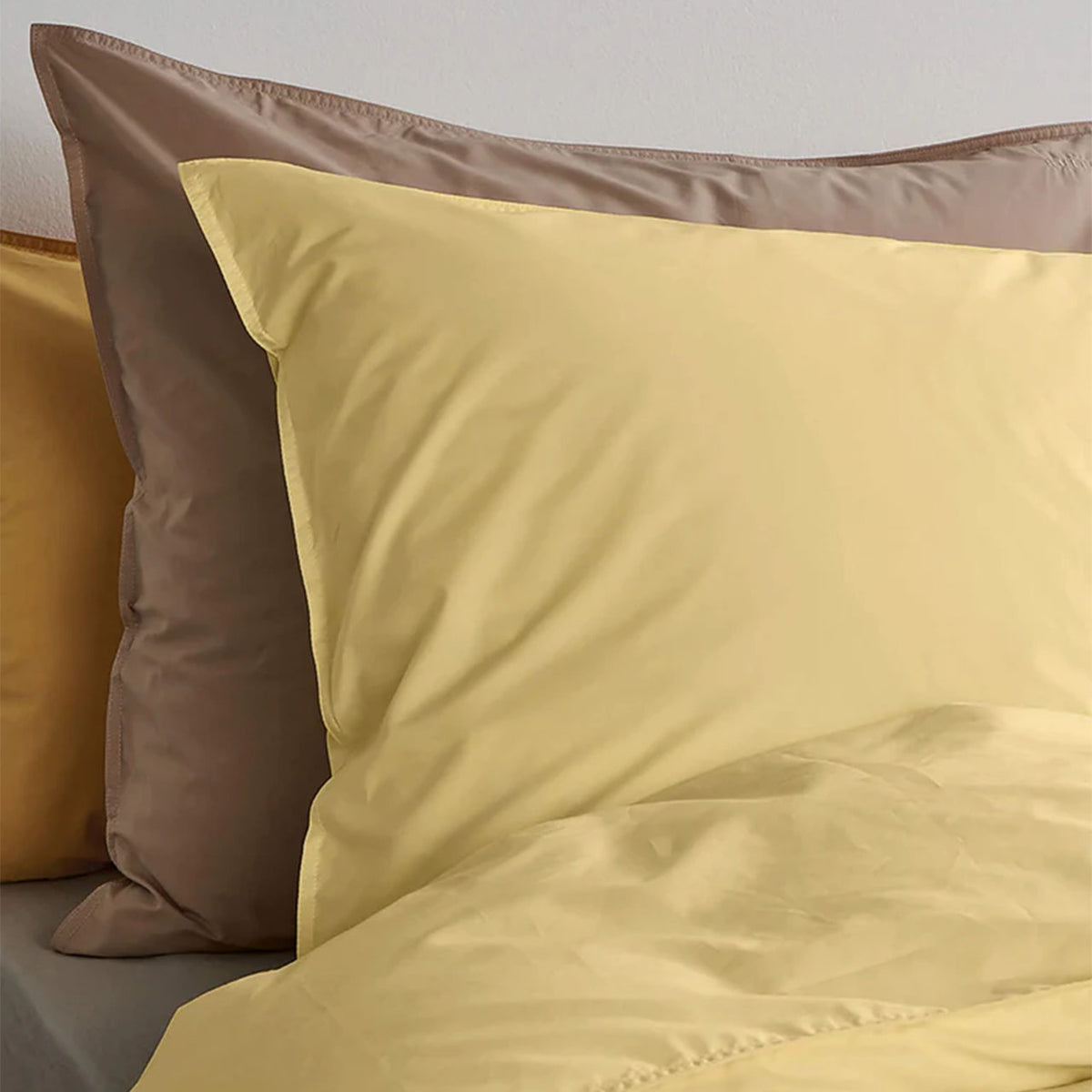 Suite702 Perkal Kissenbezug aus Bio-Baumwolle in „Butter Yellow“, davor Kissen in Braun, Nahaufnahme auf dem Bett