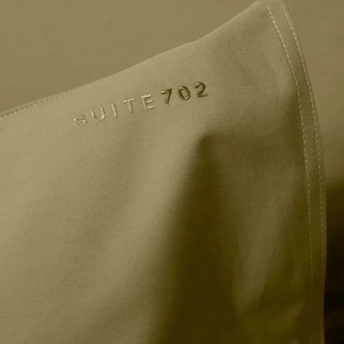 Nahaufnahme des gestickten Schriftzugs „Suite702“ auf olivgrünem Perkal-Kissenbezug
